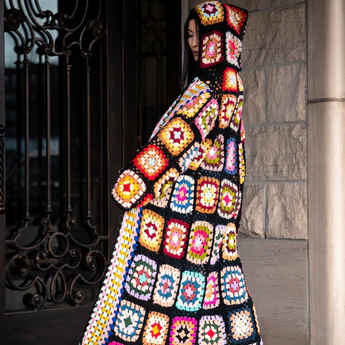 Person wearing a colorful crochet coat standing against a stone wall.