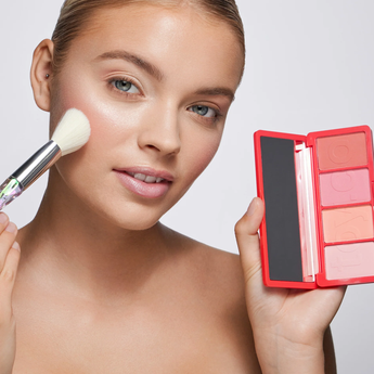 Woman applying blush with a brush and palette on a plain background