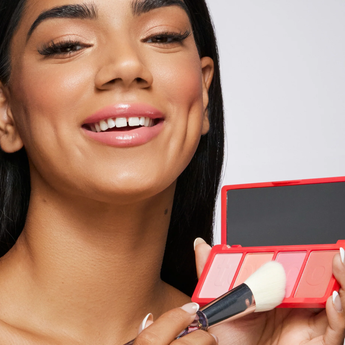 Woman holding a red makeup palette with a brush, smiling.